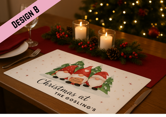 A cozy Christmas dining table set with a placemat featuring three watercolor gnomes in red hats with snowy pine trees in the background. The table is decorated with red napkins, pinecones, holly berries, and lit candles, with a softly glowing Christmas tree behind.
