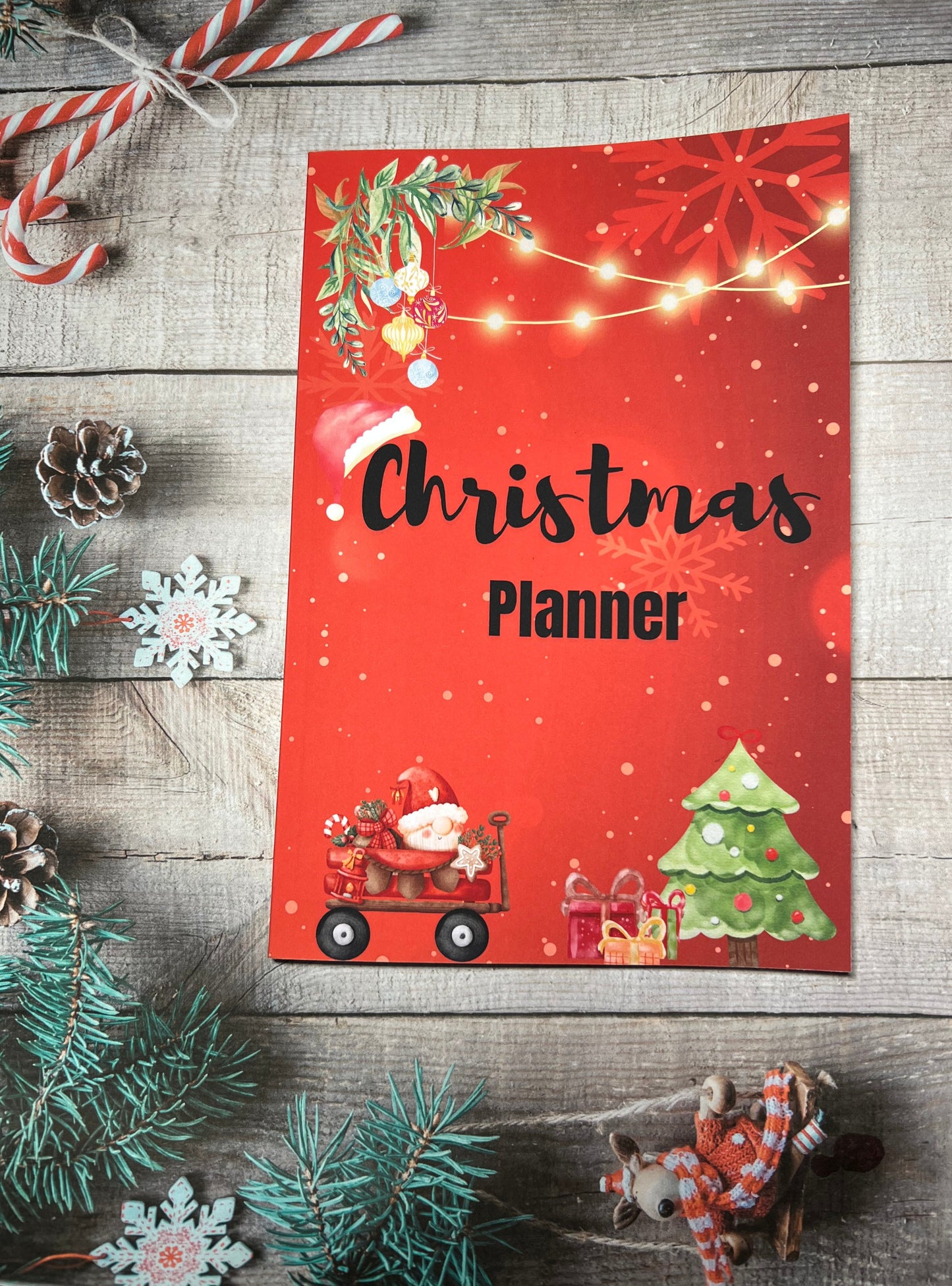 Red paperback book laying on a wooden table surrounded by Christmas decorations. The title of the book is called Christmas Planner and the cover also has pictures of Christmas tree, Presents and a Gonk dressed as Santa along the bottom with Christmas lights and decorations along the top.