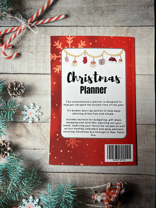 Red paperback book laying on a wooden table surrounded by Christmas decorations. This is the rear of the book which has a white box containing the title of the book being Christmas Planner and a brief description of the contents.