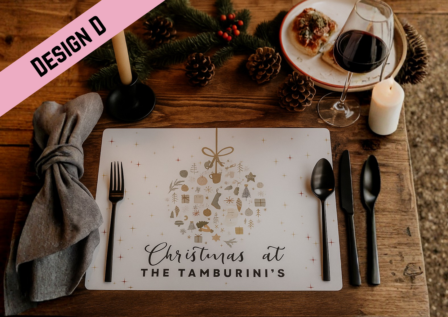 A cozy Christmas dining table is set with a personalised placemat that reads “Christmas at the Tamburrini’s.” The placemat features festive illustrations shaped like a hanging ornament, including snowflakes, trees, mittens, and gifts. The table is decorated with pine branches, pinecones, a lit candle, a plate of food, and a glass of red wine, creating a warm and inviting holiday atmosphere.