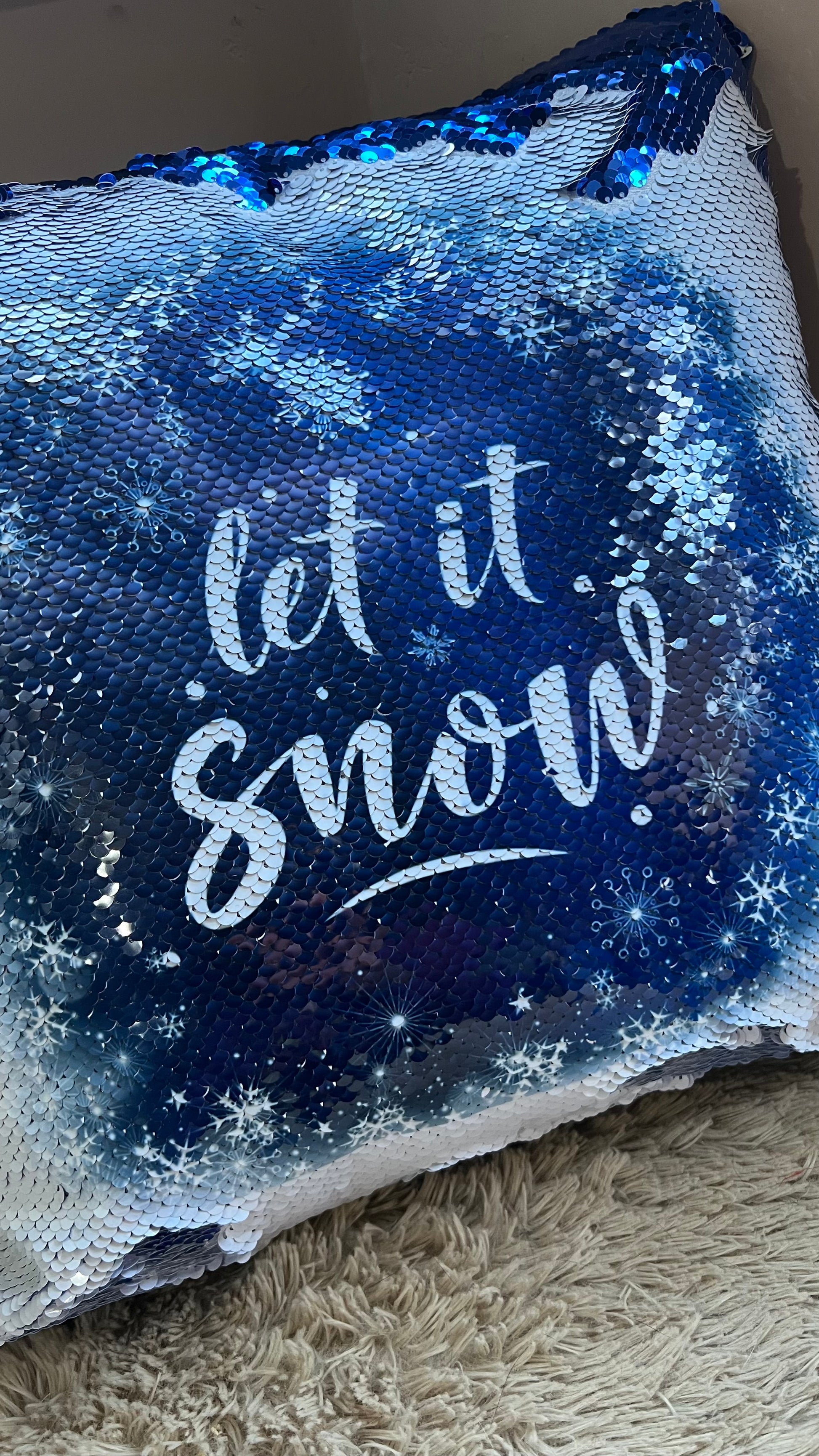 Blue sequin pillow with 'let it snow' text on a textured surface
