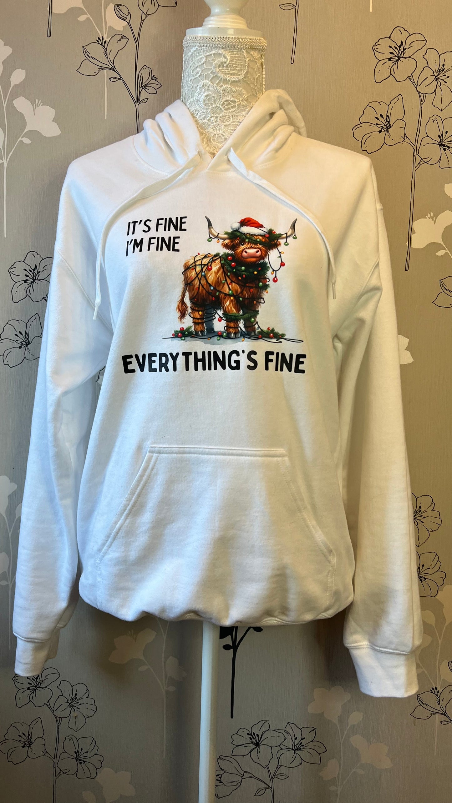 White hoodie with graphic design on a mannequin against a floral-patterned wall.