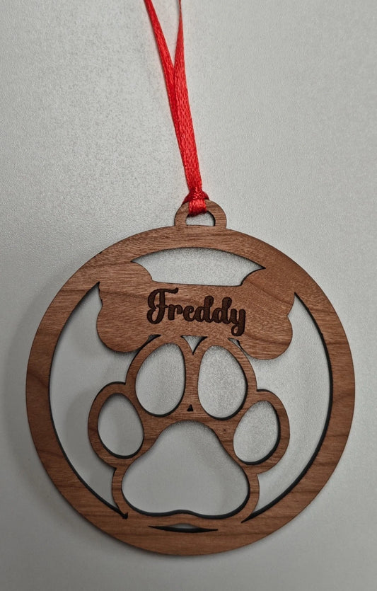 A round wooden pet ornament with a paw print and bone design, personalized with the name 'Freddy' in laser-cut lettering, hanging from a red ribbon against a neutral gray background.