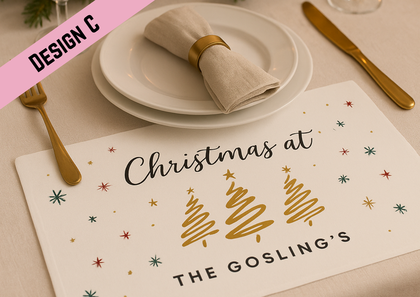 An elegant Christmas dinner setup featuring a white placemat with gold stylised Christmas trees and festive stars. The table is arranged with gold cutlery, crystal glasses, white plates, linen napkins, and candles on gold holders, with a Christmas tree twinkling in the background.
