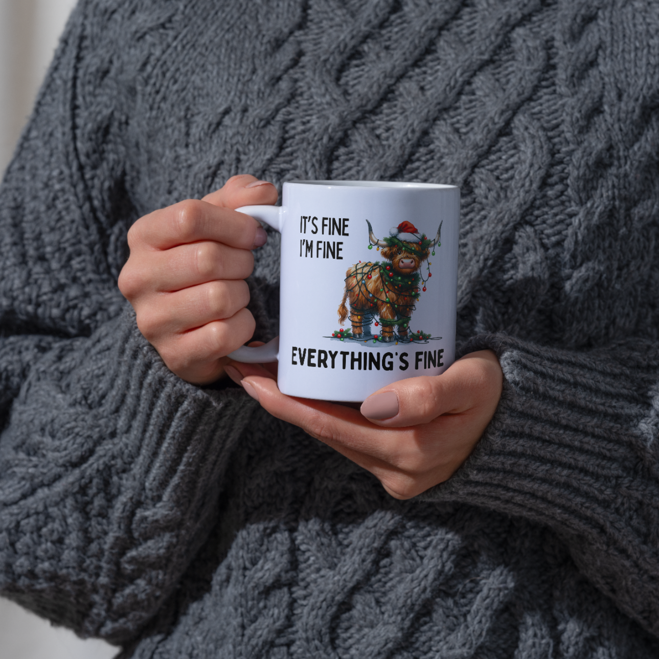 Person wearing a grey jumper holding a mug which has graphic of a cartoon highland cow wrapped in Christmas Lights with the text "It's fine I'm Fine Everything is Fine"