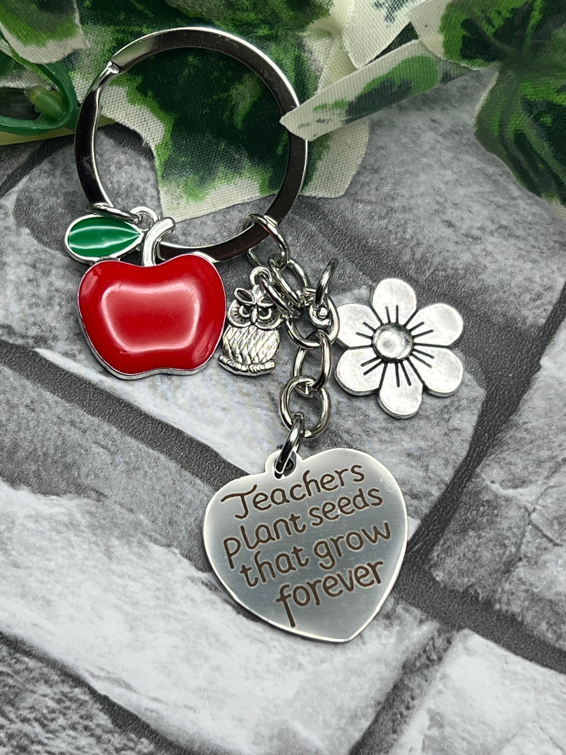 A metal king ring featuring 3 charms alongside a heart shape tag.  The tag reads "Teachers plant seeds that grow forever" and the charms are a small flower, tiny owl and a red apple.