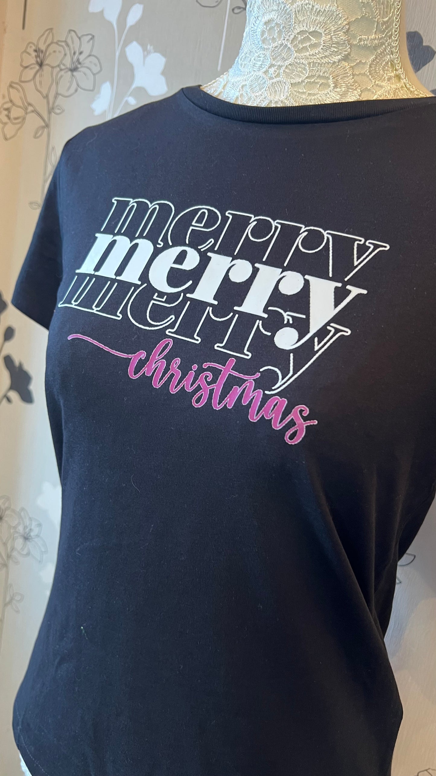 Black t-shirt with 'merry merry christmas' text on a mannequin against a floral background