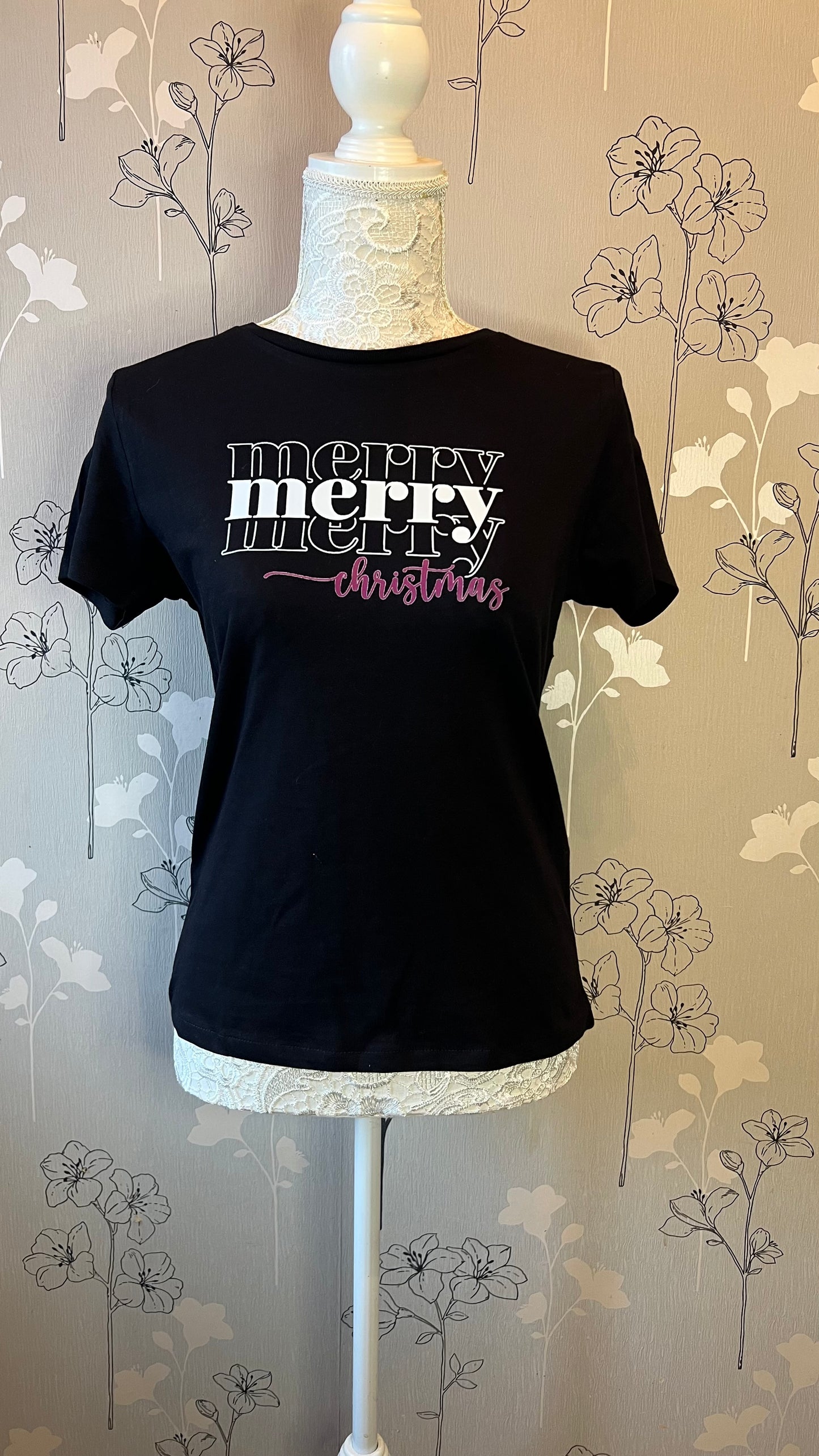 Black t-shirt with 'Merry Christmas' text on a mannequin against a floral wallpaper background