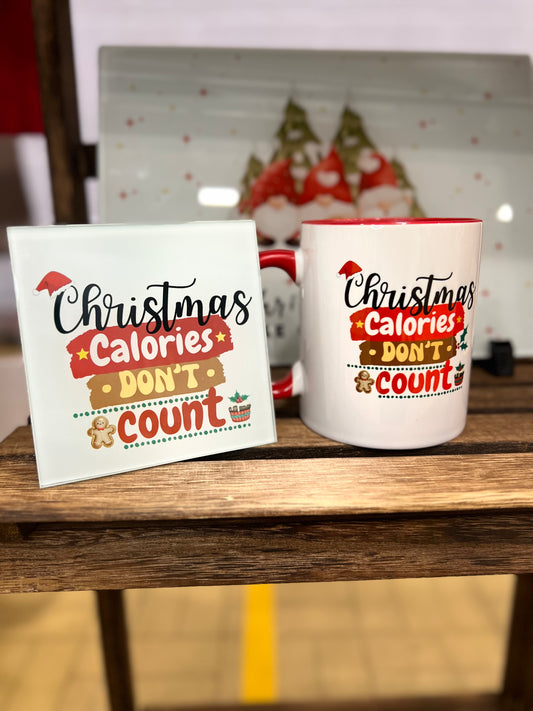 Mug and Coaster set with Christmas-themed text reading 'Christmas Calories Don't Count'. Both are standing on a wooden surface, with a blurred festive background