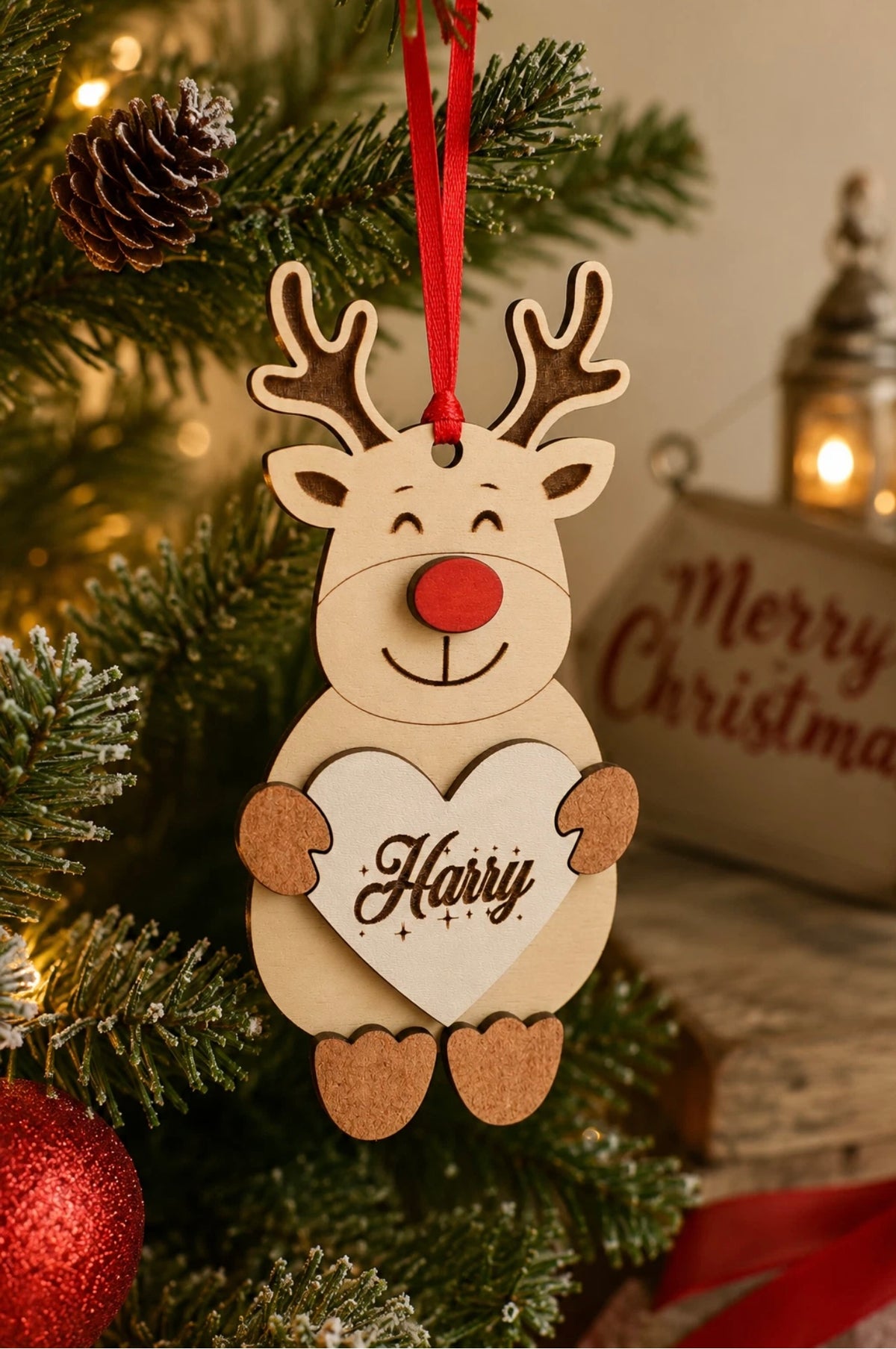 A wooden Christmas ornament shaped like a smiling reindeer with antlers, a red nose, and a red hanging ribbon, holding a heart that says “Harry.” It is displayed on a decorated Christmas tree with pinecones, warm string lights, and a blurred “Merry Christmas” sign in the background.