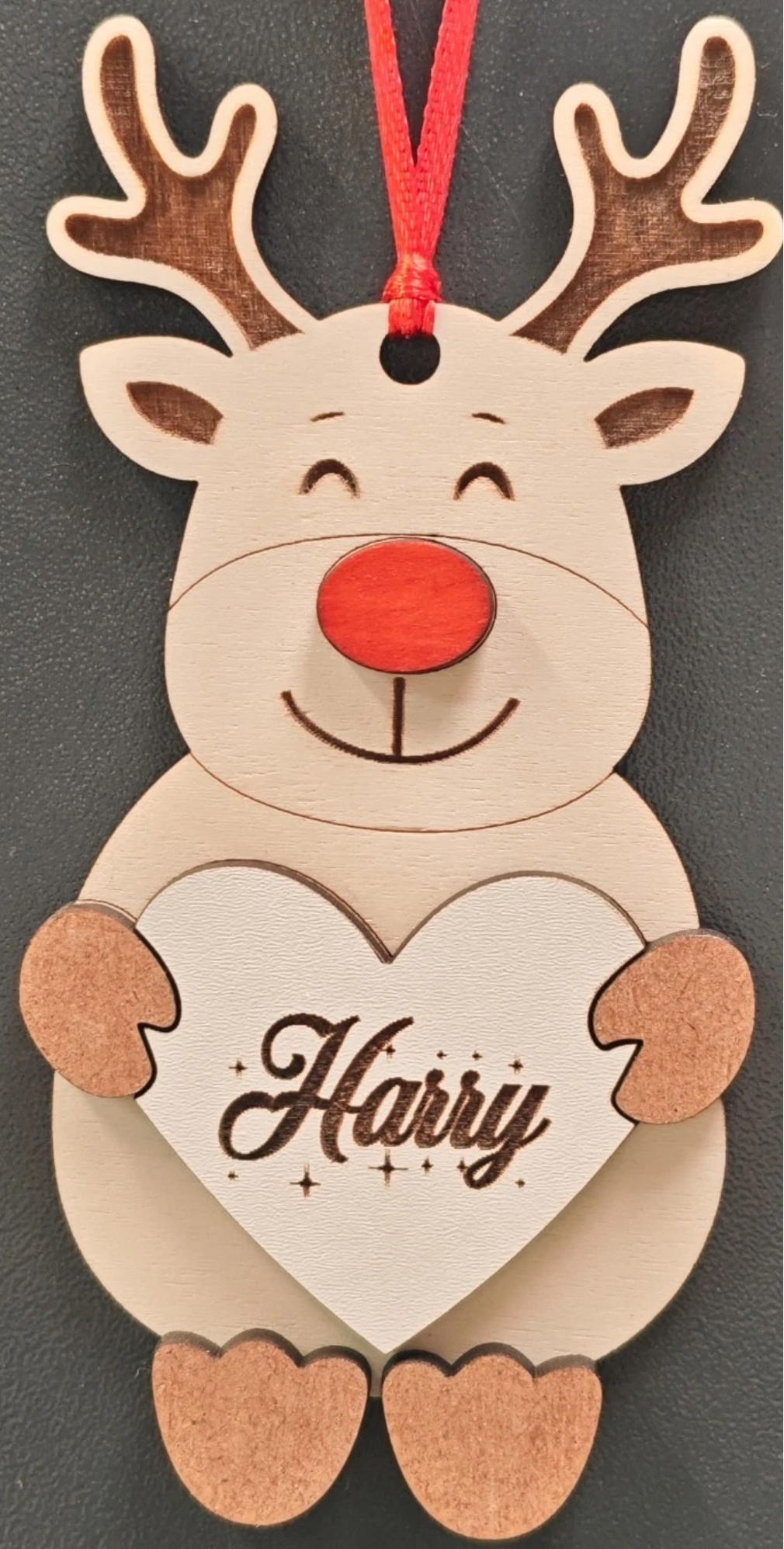 A personalized wooden Christmas ornament shaped like a smiling reindeer holding a heart with the name 'Harry' engraved, hanging from a red ribbon against a dark gray background.