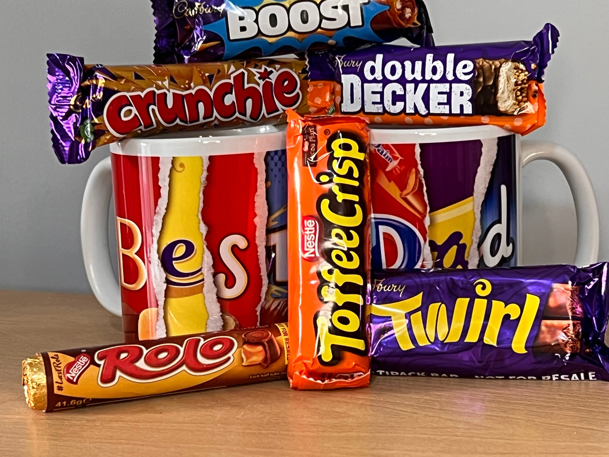 2 (two) mugs side by side containing the words Best Dad which are formed using letters from images on chocolate wrappers.  The mugs are surrounded by bars of chocolate